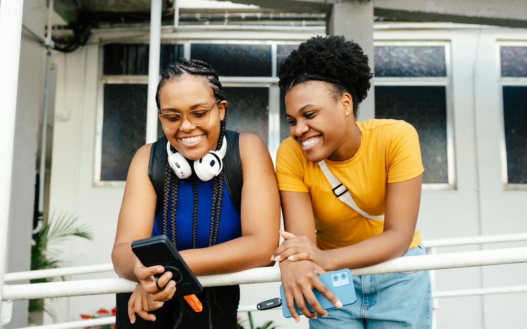 Duas profissionais fora do escritório conferindo o celular, uma delas com fones no pescoço